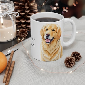 Pode incluir: Caneca de café de cerâmica branca com uma ilustração de golden retriever e o texto "Golden's Forever". A caneca está cheia de café e está sobre uma superfície branca com uma pinha marrom e um pau de canela.