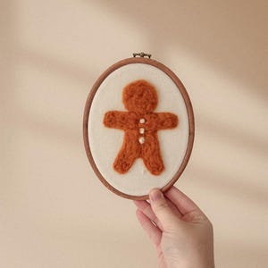 May include: An oval-shaped, wooden embroidery hoop holds a felt gingerbread man. The gingerbread man is a warm orange color with three white button accents. The background is a cream-colored fabric. The hoop is held by a hand.