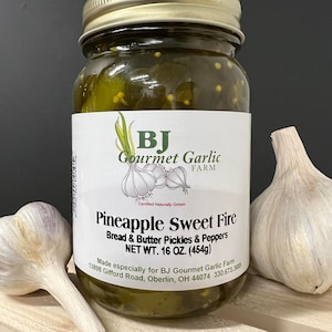 May include: A glass jar filled with green bread and butter pickles and peppers. The jar has a gold lid and a white label that reads "BJ Gourmet Garlic Farm" and "Pineapple Sweet Fire". The label also includes the text "Certified Naturally Grown", "Made especially for BJ Gourmet Garlic Farm", and "13898 Gifford Road, Oberlin, OH 44074 330.673.3689".