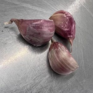 May include: Three purple garlic cloves on a metallic surface. The cloves have a layered texture and a slight curve, with a light-colored tip. The image is a close-up, highlighting the details of the garlic.