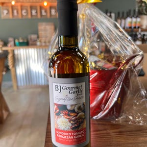 May include: A bottle of BJ Gourmet Garlic Farm Signature Sauce. The label features images of sundried tomatoes, parmesan cheese, and garlic. The bottle is filled with a golden liquid and topped with a black cap. The text "SUNDRIED TOMATO PARMESAN GARLIC" is visible.