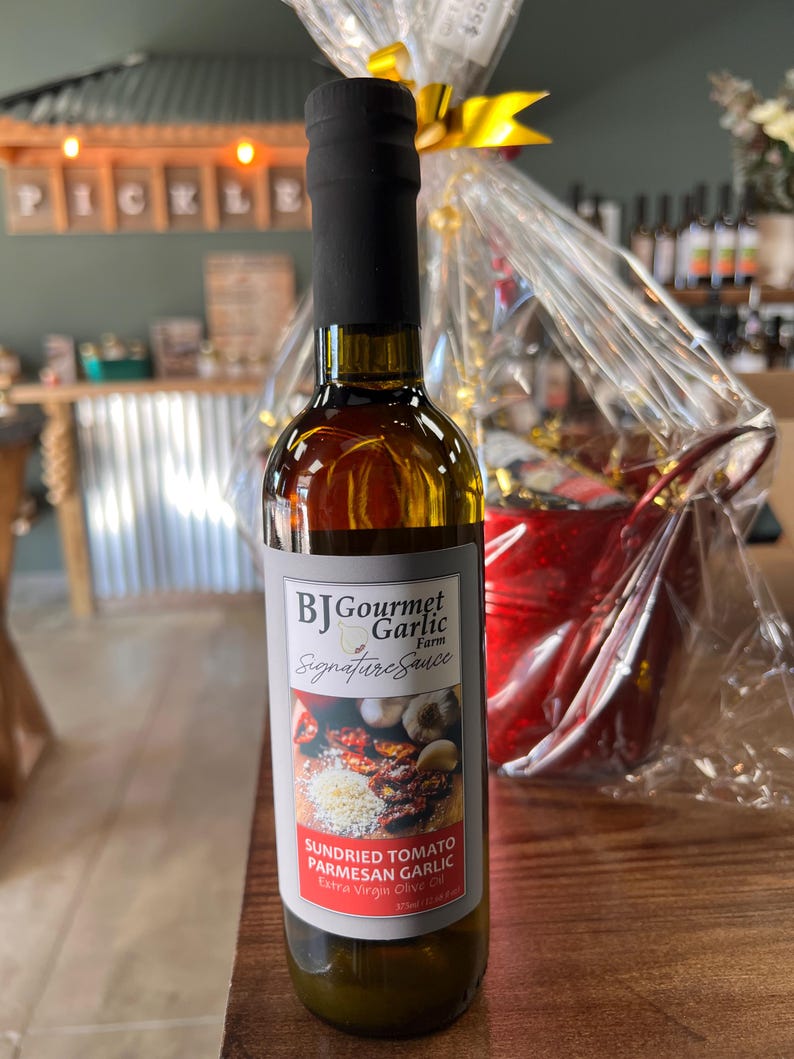 May include: A bottle of BJ Gourmet Garlic Farm Signature Sauce. The label features images of sundried tomatoes, parmesan cheese, and garlic. The bottle is filled with a golden liquid and topped with a black cap. The text "SUNDRIED TOMATO PARMESAN GARLIC" is visible.