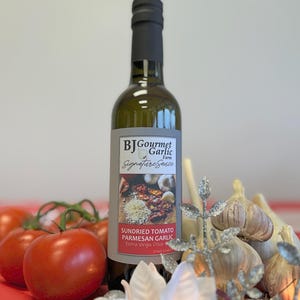 May include: A bottle of BJ Gourmet Garlic Farm Signature Sauce, featuring sundried tomato, Parmesan garlic, and extra virgin olive oil. The bottle is surrounded by fresh tomatoes, garlic bulbs, and decorative accents.