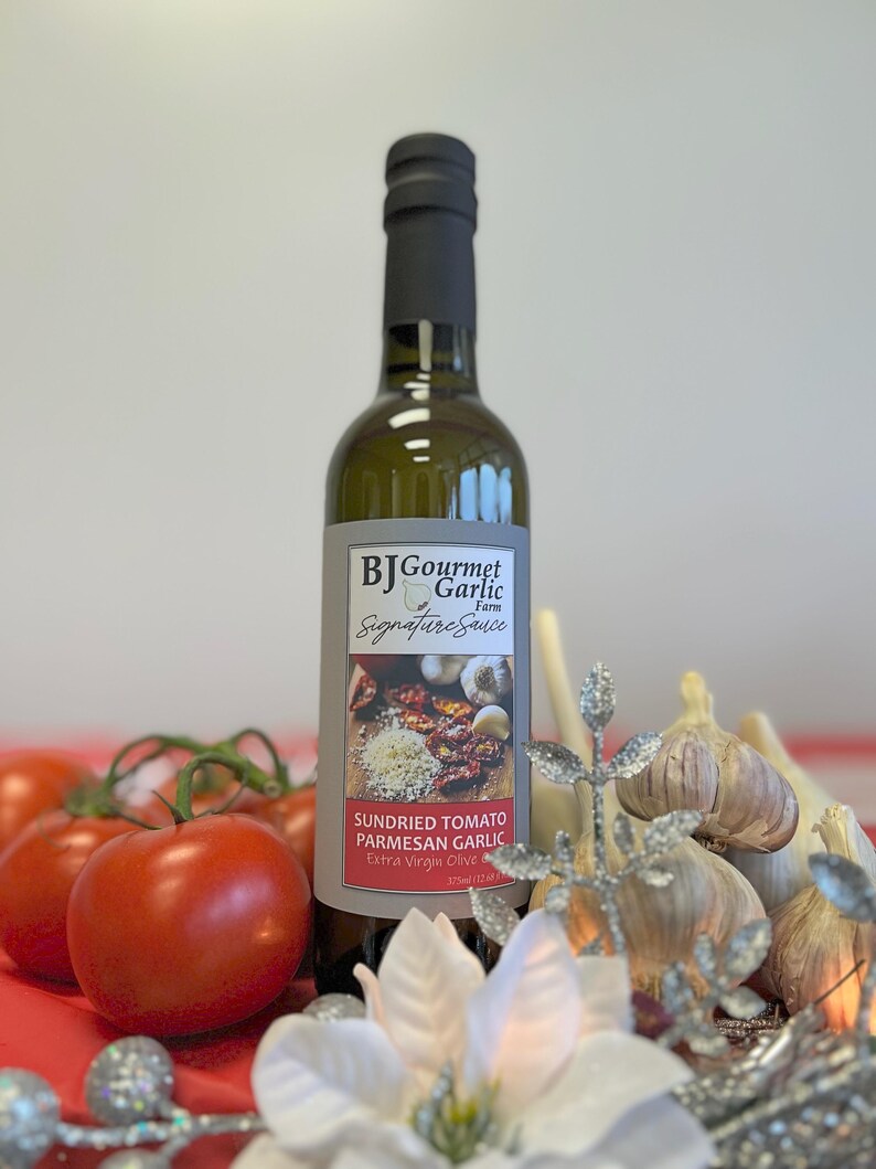 May include: A bottle of BJ Gourmet Garlic Farm Signature Sauce, featuring sundried tomato, Parmesan garlic, and extra virgin olive oil. The bottle is surrounded by fresh tomatoes, garlic bulbs, and decorative accents.