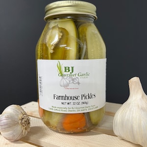 May include: A glass jar filled with green dill pickle spears and a few carrot slices. The jar has a gold lid and a white label that reads "BJ Gourmet Garlic Farm" and "Farmhouse Pickles". The label also states "Net Wt. 32 oz. (908g)" and includes the farm's address and phone number.