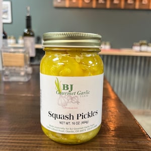 May include: A jar of squash pickles with a gold lid and a white label. The label reads "BJ Gourmet Garlic Farm" and "Squash Pickles, NET WT. 16 OZ. (454g)". The jar is filled with yellow pickles and sits on a wooden surface.