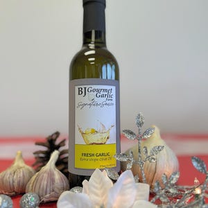 K&ouml;nnte beinhalten: Eine Glasflasche mit frischem Knoblauch-Oliven&ouml;l extra vergine mit einem wei&szlig;en Etikett, auf dem "BJ Gourmet Garlic Farm Signature Sauce" und "Fresh Garlic Extra Virgin Olive Oil 375ml (12.68 fl oz)" steht. Die Flasche steht auf einer roten Oberfl&auml;che mit einer wei&szlig;en Blume, Tannenzapfen und Knoblauchzehen.