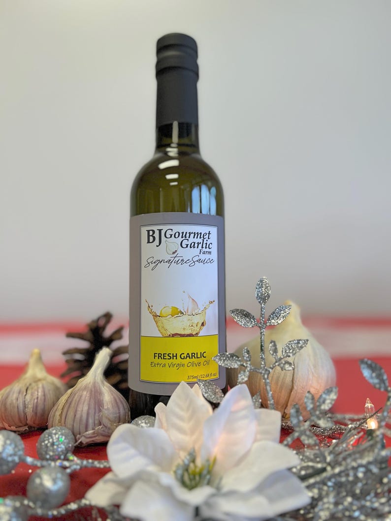 K&ouml;nnte beinhalten: Eine Glasflasche mit frischem Knoblauch-Oliven&ouml;l extra vergine mit einem wei&szlig;en Etikett, auf dem "BJ Gourmet Garlic Farm Signature Sauce" und "Fresh Garlic Extra Virgin Olive Oil 375ml (12.68 fl oz)" steht. Die Flasche steht auf einer roten Oberfl&auml;che mit einer wei&szlig;en Blume, Tannenzapfen und Knoblauchzehen.