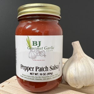 May include: A glass jar of red pepper patch salsa with a gold lid. The jar is labeled "BJ Gourmet Garlic Farm" and "Pepper Patch Salsa". The jar is sitting on a wooden surface next to a large garlic clove.