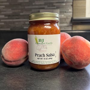 May include: A jar of peach salsa with a gold lid and a white label featuring the text "Peach Salsa". Two ripe peaches flank the jar. The label also includes the text "BJ Gourmet Garlic Farm".