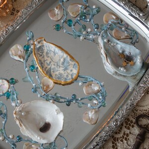 May include: A silver tray with ornate edges holds decorative oyster shells, beaded strands, and small jewelry. The shells are white and blue, some with gold trim. The beaded strands are blue and white, with pearls and glass beads.