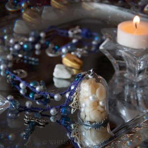May include: A decorative display featuring a lit candle in a glass holder, a glass dome filled with white objects, and a beaded necklace with blue and white beads. The scene is set on a reflective surface.