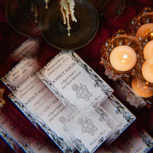Puede incluir: Tres folletos de papel blanco con el título "Witchey & Wool - A Pocket Grimoire - Vol. 1" y "Poppet Magic: A Quick Guide to Doll-Based Spellwork" en la portada. Los folletos están apilados uno encima del otro y están rodeados de velas.