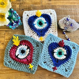 May include: Three crocheted squares with a heart-shaped evil eye design. The squares are in shades of blue, white, red, and yellow.