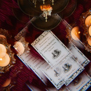 Op de afbeelding: Een close-up van een wit boekje met zwarte tekst en een zwart-witte illustratie van een theekopje. Het boekje is getiteld "TASSEOGRAPHY: A Quick Guide to Reading Tea Leaves" en maakt deel uit van een "Witchy & Wool" pakket.