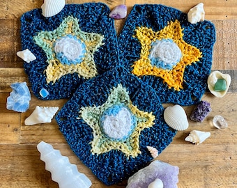 May include: Three crocheted star-shaped squares in shades of blue, green, and yellow. The squares are arranged in a triangle shape and surrounded by various small stones and seashells.