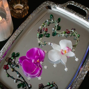 May include: A silver tray with ornate handles holds two orchid flower hair accessories, one purple and one white with pink accents. Decorative beaded vines and candles in holders surround the tray, creating a romantic ambiance.