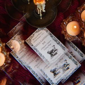 Puede incluir: Una pila de pequeños libros blancos con texto negro e ilustraciones. Los libros se titulan "Spell Casting 101: A Beginner's Guide to Crafting Magic" y presentan el texto "Witchy & Wool: A Pocket Grimoire - Vol. 1".