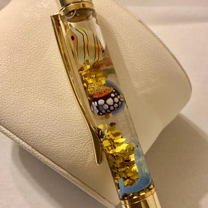May include: A clear liquid-filled pen with gold-colored metal accents. The barrel contains floating gold glitter and small, colorful illustrations of various tropical fish, including a yellow striped fish and a spotted fish, along with a blue dolphin.