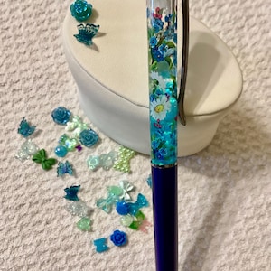 May include: A decorative pen with a clear barrel filled with blue and white floral designs. The pen has a dark blue body and a silver clip and tip. Scattered around the pen are small, decorative blue and green embellishments.