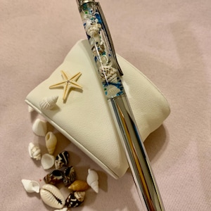May include: A silver pen with a clear barrel filled with seashells, blue accents, and glitter. A starfish and various seashells are arranged near the pen on a white square pillow. The pen is a unique writing instrument.