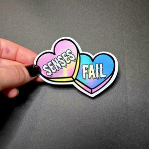 May include: A sticker featuring two heart-shaped designs. One heart is pink and yellow with the word "SENSES" in white, and the other is blue with the word "FAIL" in white. The sticker has a holographic effect.