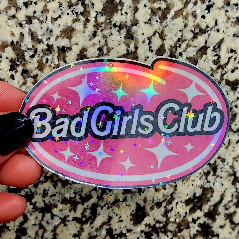 Op de afbeelding: Ovale sticker met een roze achtergrond, witte rand en de tekst "Bad Girls Club" in zwart en wit. De sticker heeft een holografische, iriserende afwerking met stervormige accenten.