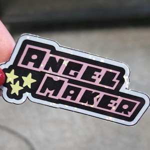 May include: A rectangular sticker with the text "ANGEL MAKER" in pink block letters outlined in black. The sticker has a black background and a white border with three yellow stars on the left side.