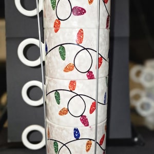 Pode incluir: Uma pilha de quatro canecas de cerâmica branca com um design de luzes de Natal em um suporte de metal prateado. Cada caneca apresenta uma sequência de lâmpadas coloridas e brilhantes. As canecas têm alças brancas.