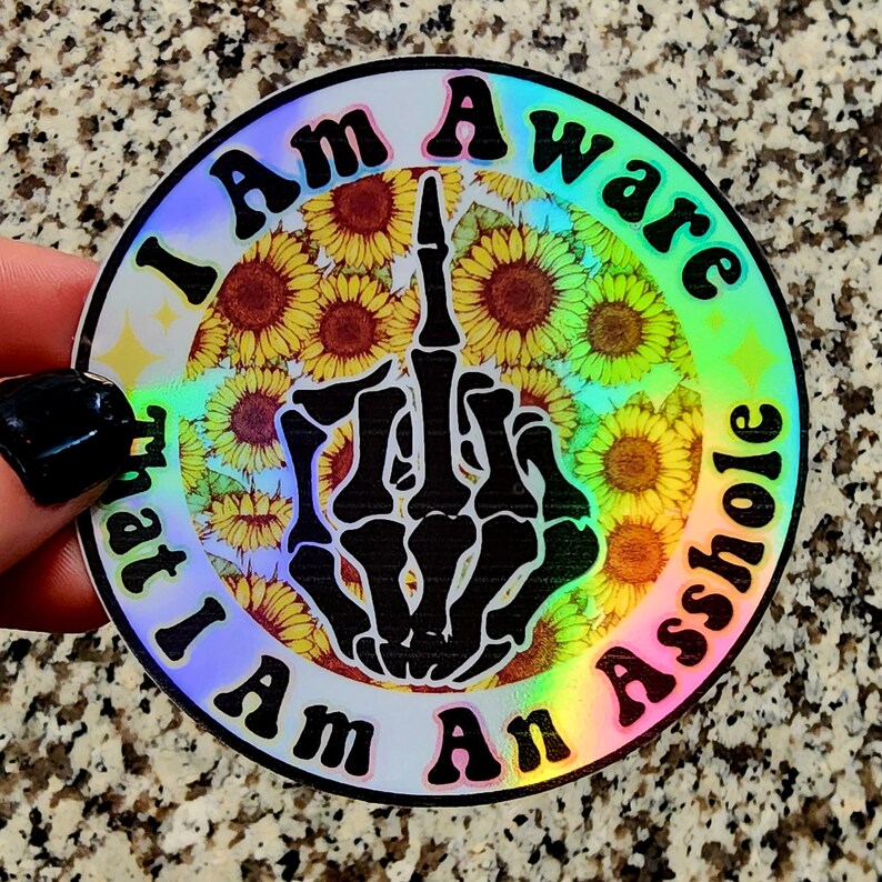 May include: A round sticker with a holographic finish. The sticker features a black skeleton hand giving the middle finger, surrounded by sunflowers. The text "I Am Aware That I Am An Asshole" is printed around the edge in black.