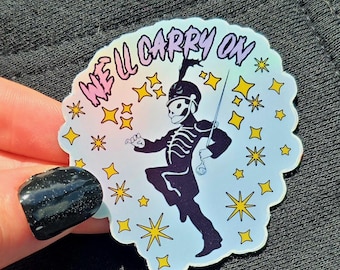 May include: A sticker featuring a marching band skeleton in a black uniform, holding a sword. The sticker has a light blue background with yellow stars and the text "WE'LL CARRY ON" in pink and purple. The sticker is held by a hand with a black glitter nail.