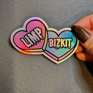 May include: A holographic sticker featuring two connected heart shapes. The left heart is pink and reads "LIMP" in dripping black letters. The right heart is blue and reads "BIZKIT" in dripping black letters. The sticker has a rainbow sheen.