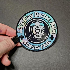 Könnte beinhalten: Ein runder Aufkleber mit holografischem Finish. Der Aufkleber zeigt einen Schädel mit medizinischem Stirnband, mit dem Text "Let's Play Doctor, Babe" und "Senses Fail" in einem kreisförmigen Design. Schwarze Pluszeichen befinden sich auf beiden Seiten.