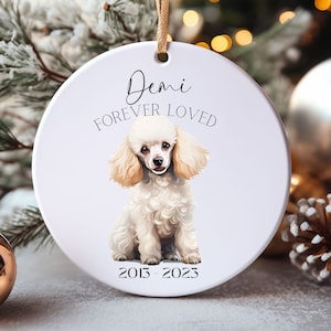 May include: White ceramic Christmas ornament with a watercolor illustration of a poodle. The ornament has the text "Demi Forever Loved" and the years "2013-2023". A gold ribbon is attached for hanging. The ornament is surrounded by holiday decorations.