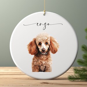 May include: White ceramic Christmas ornament with a brown and white poodle dog illustration and the name "Enzo" written in cursive.