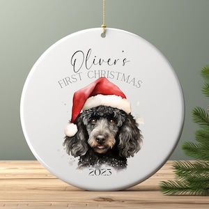 May include: A white ceramic Christmas ornament featuring a black poodle wearing a Santa hat. The text "Oliver's First Christmas 2023" is written on the ornament. A gold string is attached for hanging.