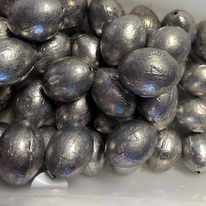 May include: A pile of silver-colored, egg-shaped fishing weights. Each weight has a small hole for attaching fishing line. The weights are smooth and metallic, with a slightly textured surface. They are likely made of lead or a similar dense metal.
