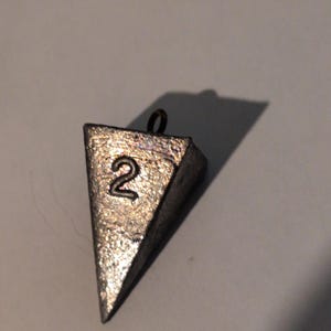 May include: A triangular, silver-colored fishing weight with the number "2" embossed on its surface. The weight has a small loop at the top for attaching to fishing line. The weight is designed to sink fishing lures or bait.