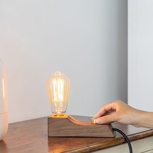 May include: A wooden lamp base with a vintage-style light bulb. The lamp has a brass switch and is turned on. The lamp is on a wooden surface next to a white vase.