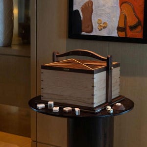 May include: A wooden box with a handle and a geometric design sits on a small, dark round table. Dice and game tiles are scattered around the box. A painting with a foot and sandals hangs in the background.