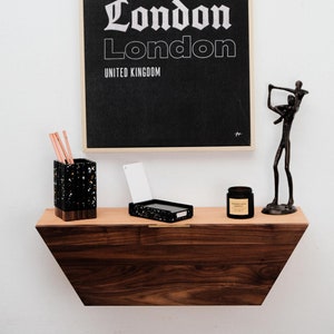 May include: A black and white poster with the text "LONDON London London UNITED KINGDOM" hanging on a white wall above a wooden shelf. The shelf has a black and white speckled pencil holder, a white card, a black and white speckled box, a small glass jar with a label, and a black metal sculpture of a person holding another person.
