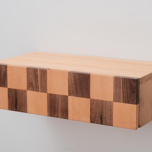 Op de afbeelding: Een rechthoekige zwevende houten plank met een dambordpatroon op de voorrand. De afwisselende vierkanten zijn licht en donker van kleur. De plank is aan een witte muur bevestigd.