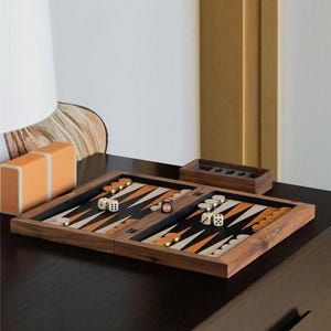 May include: A wooden backgammon set with a black playing surface, orange and white triangles, and wooden game pieces. Dice are scattered across the board. A small wooden box and an orange box are also visible.