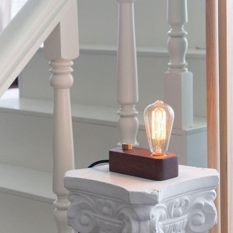 May include: A small wooden table lamp with a vintage-style light bulb. The lamp is on a white pedestal and has a brown wooden base.