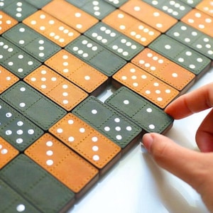 Puede incluir: Primer plano de un juego de dominó en progreso. Los dominós están dispuestos en un patrón de cuadrícula, con colores alternos verde oliva y marrón claro. Cada dominó tiene puntos blancos. Una mano alcanza un dominó.