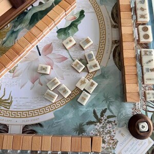 May include: A Mahjong game set with wooden tiles and racks, scattered on a floral patterned game board. The tiles are off-white with detailed symbols. A wooden dice cup and dice are also visible.