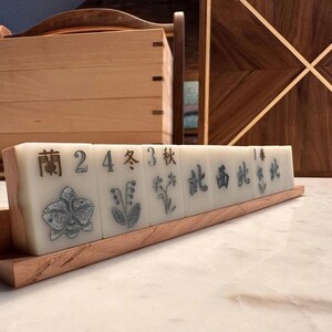 May include: A set of ivory-colored Mahjong tiles with black Chinese characters and floral illustrations, displayed on a wooden stand. The tiles are arranged in a row, showcasing the intricate designs and craftsmanship.