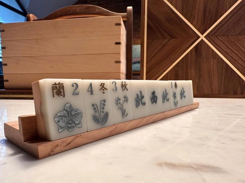 May include: A set of ivory-colored Mahjong tiles with black Chinese characters and floral illustrations, displayed on a wooden stand. The tiles are arranged in a row, showcasing the intricate designs and craftsmanship.