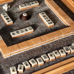 May include: A wooden Mahjong set with ivory tiles featuring black symbols and dots. The set includes dice and a wooden tile holder. The tiles are arranged on a wooden surface, showcasing the game's components.