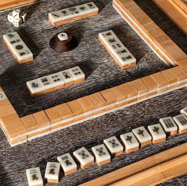 May include: A wooden Mahjong set with ivory tiles featuring black symbols and dots. The set includes dice and a wooden tile holder. The tiles are arranged on a wooden surface, showcasing the game's components.